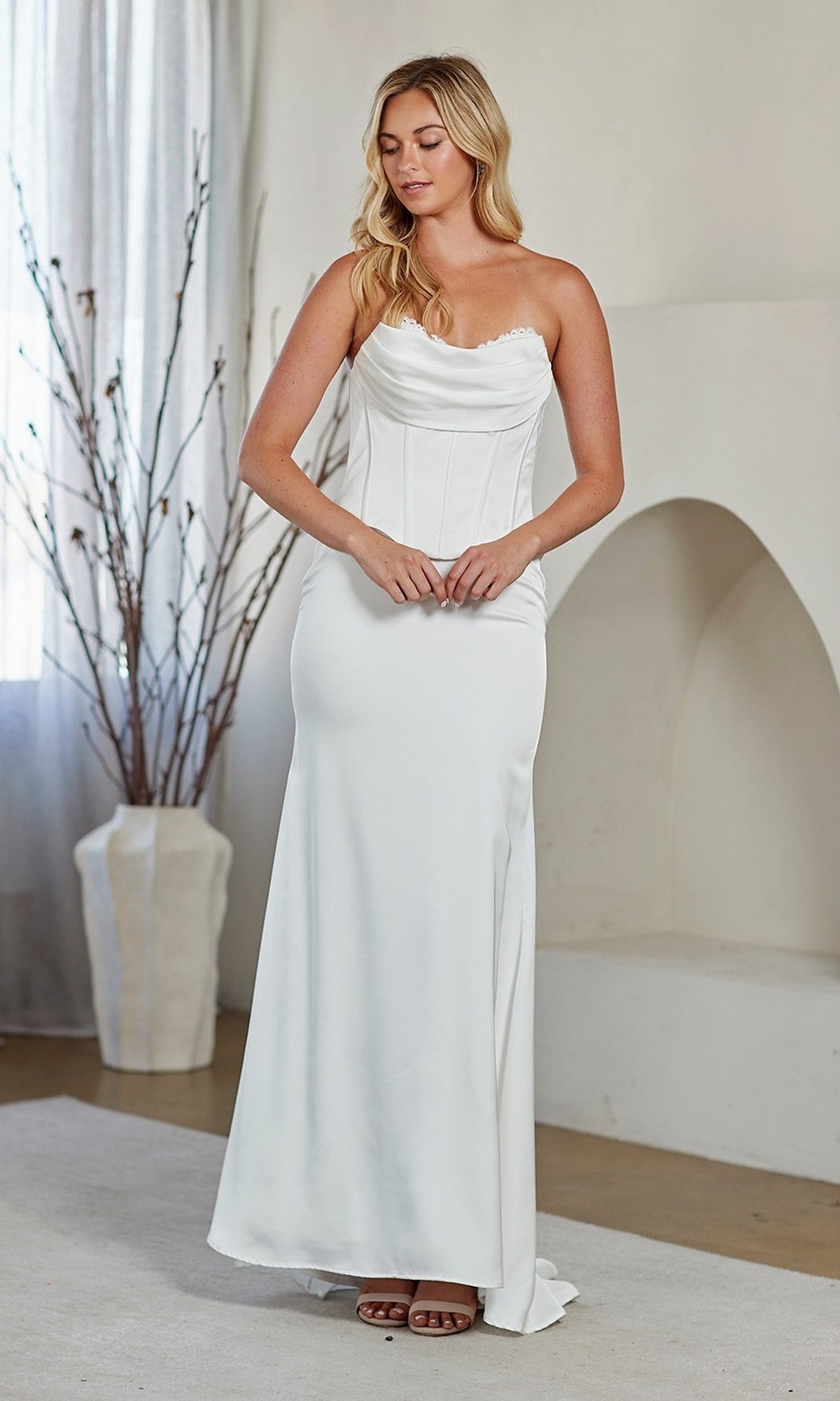 A woman stands indoors wearing the Amelia Couture 6137 long prom dress, featuring a strapless fitted corset bodice and flowing skirt. A vase with branches and a fireplace are visible in the background.